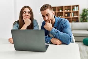 Young caucasian couple working using computer laptop at home serious face thinking about question with hand on chin, thoughtful about confusing idea