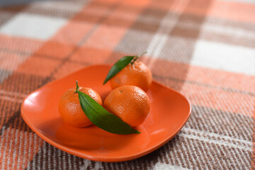 Three mandarins with green leaves on an orange plate on a checked wool plaid. Top view. Diet....