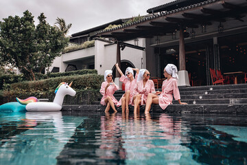 Four adorable skinnu and lovely female models in identical clothes posing and sitting next to the pool with inflatable toys against the backdrop of the villa and houses. Chill out concept