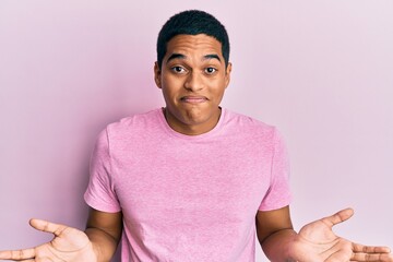 Young handsome hispanic man wearing casual pink t shirt clueless and confused expression with arms...