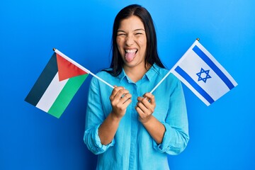 Young latin woman holding palestine and israel flags sticking tongue out happy with funny...