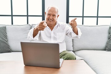 Senior man using laptop at home sitting on the sofa pointing fingers to camera with happy and funny face. good energy and vibes.