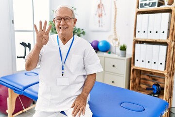 Obraz premium Senior physiotherapy man working at pain recovery clinic showing and pointing up with fingers number four while smiling confident and happy.