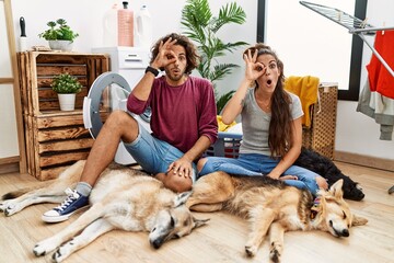 Young hispanic couple doing laundry with dogs doing ok gesture shocked with surprised face, eye looking through fingers. unbelieving expression.