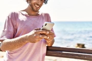 Young hispanic man smiling happy using smartphone standing at the beach.