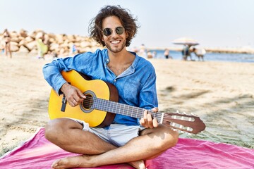 Young hispanic man playing classical guitar sitting on sand at the beach.