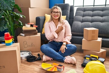Young hispanic woman moving to a new home sitting on the floor cheerful with a smile on face...
