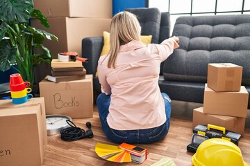Young hispanic woman moving to a new home sitting on the floor posing backwards pointing ahead with finger hand