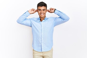 Young hispanic man wearing business shirt standing over isolated background smiling pulling ears...