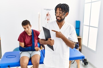 Young hispanic man working at pain recovery clinic with a man with broken arm pointing aside worried and nervous with forefinger, concerned and surprised expression