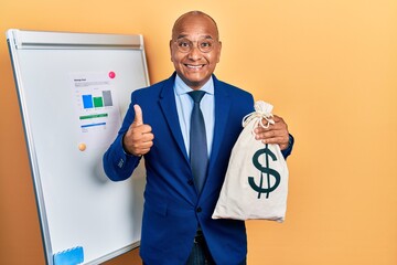 Middle age latin man wearing business suit holding dollars bag smiling happy and positive, thumb up...