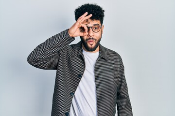 Young arab man with beard wearing glasses doing ok gesture shocked with surprised face, eye looking through fingers. unbelieving expression.