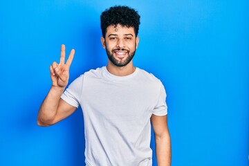 Naklejka premium Young arab man with beard wearing casual white t shirt smiling looking to the camera showing fingers doing victory sign. number two.