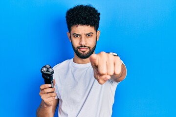 Young arab man with beard holding electric razor machine pointing with finger to the camera and to...