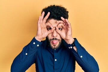 Young arab man with beard wearing casual shirt doing ok gesture like binoculars sticking tongue out, eyes looking through fingers. crazy expression.