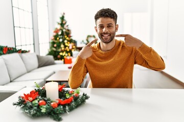 Arab young man sitting on the table by christmas tree smiling cheerful showing and pointing with fingers teeth and mouth. dental health concept.