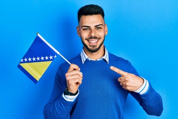 Young hispanic man with beard holding bosnia herzegovina flag smiling happy pointing with hand and finger