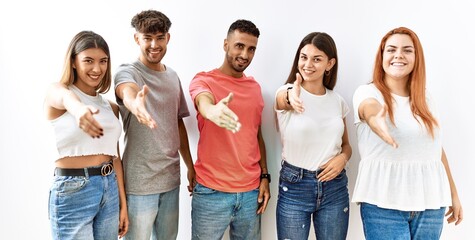 Group of young friends standing together over isolated background smiling friendly offering handshake as greeting and welcoming. successful business.