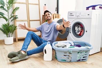 Young hispanic man putting dirty laundry into washing machine looking at the camera smiling with open arms for hug. cheerful expression embracing happiness.
