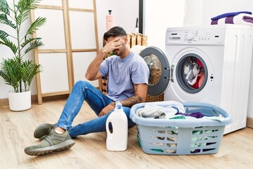 Young hispanic man putting dirty laundry into washing machine covering eyes with hand, looking serious and sad. sightless, hiding and rejection concept