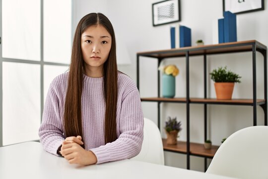 Young chinese girl wearing casual clothes sitting on the table at home with serious expression on face. simple and natural looking at the camera.