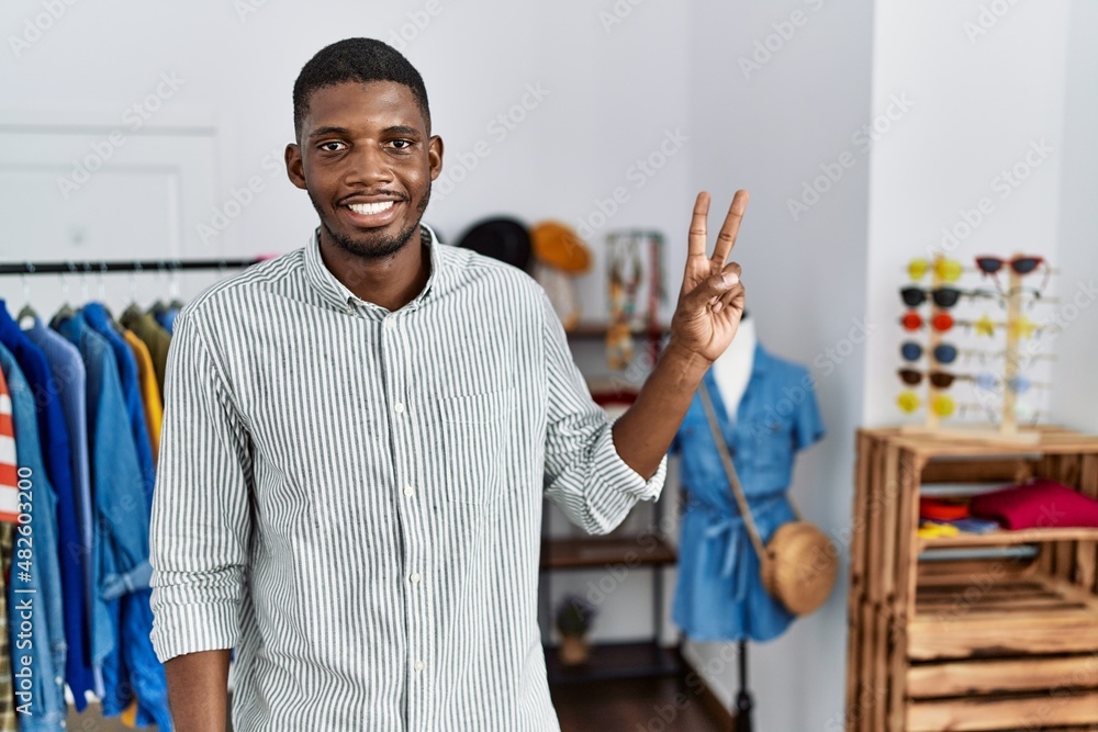 Poster Young african american man working as manager at retail boutique smiling with happy face winking at the camera doing victory sign. number two.