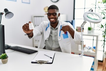 Naklejka na ściany i meble Young african american doctor man wearing doctor uniform holding prescription pills at the clinic pointing finger to one self smiling happy and proud