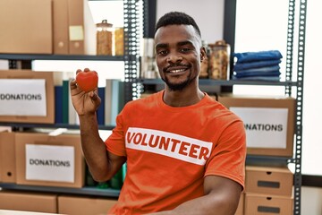 Young african man wearing volunteer t shirt at donations stand holding heart looking positive and...