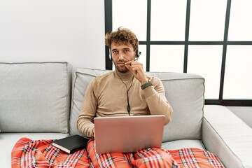 Young handsome man with beard wearing operator headset working from home mouth and lips shut as zip...