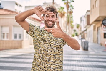 Young hispanic man standing at the street smiling making frame with hands and fingers with happy...