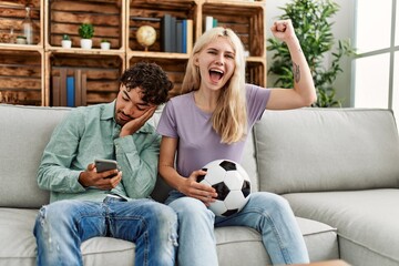 Woman smiling happy watching soccer match and boyfriend boring using smartphone at home.