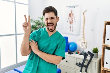 Young man with beard working at pain recovery clinic smiling with happy face winking at the camera doing victory sign. number two.