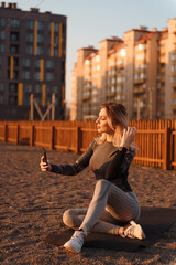 Sporty young woman sitting on yoga mat near playground, holding cellphone in her hand and doing selfie. The girl enjoys the sunset after an active workout.