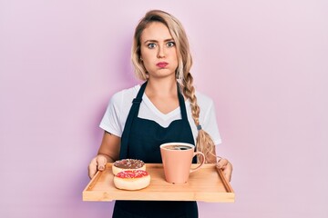 Beautiful young blonde woman wearing waitress apron holding breakfast tray puffing cheeks with funny face. mouth inflated with air, catching air.