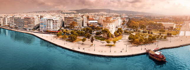Aerial scenic panorama of the main symbol of Thessaloniki city - the White Tower with boat tour ship at the pier. Concept of travel landmarks in Greece and urban development.