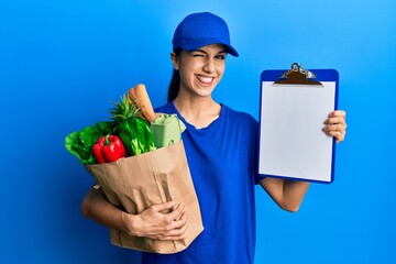 Young hispanic woman wearing courier uniform with groceries from supermarket and clipboard winking looking at the camera with sexy expression, cheerful and happy face.