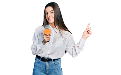Young brunette teenager holding reporter microphone smiling happy pointing with hand and finger to...
