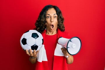 Middle age hispanic woman football hooligan holding ball and using megaphone afraid and shocked with surprise and amazed expression, fear and excited face.