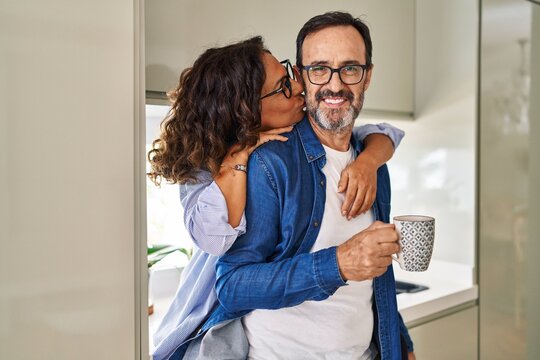 Middle age hispanic couple kissing and hugging each other drinking coffee at kitchen