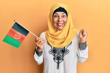 Middle age hispanic woman holding afghanistan flag screaming proud, celebrating victory and success...