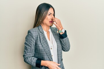 Young hispanic woman wearing business clothes smelling something stinky and disgusting, intolerable smell, holding breath with fingers on nose. bad smell