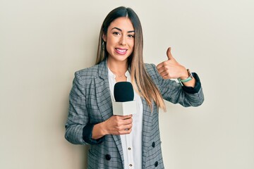 Young hispanic woman holding reporter microphone smiling happy and positive, thumb up doing...