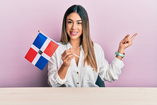 Young hispanic woman holding dominican republic flag sitting on the table smiling happy pointing with hand and finger to the side