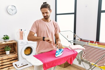 Young hispanic man ironing clothes at home checking the time on wrist watch, relaxed and confident