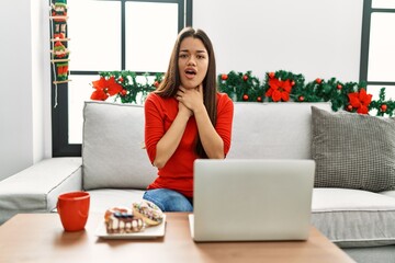 Obraz na płótnie Canvas Young brunette woman using laptop sitting on the sofa on christmas shouting and suffocate because painful strangle. health problem. asphyxiate and suicide concept.