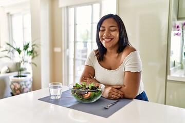 Obraz na płótnie Canvas Young hispanic woman eating healthy salad at home excited for success with arms raised and eyes closed celebrating victory smiling. winner concept.