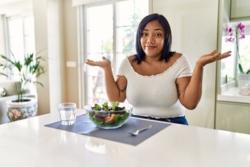Obraz na płótnie Canvas Young hispanic woman eating healthy salad at home clueless and confused expression with arms and hands raised. doubt concept.