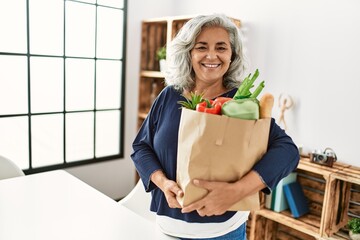 Middle age grey-haired woman holding paper bag with groceries standing at home.