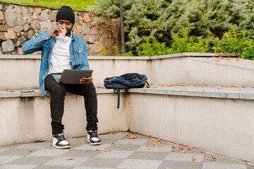 Black young man working with laptop while drinking coffee on bench
