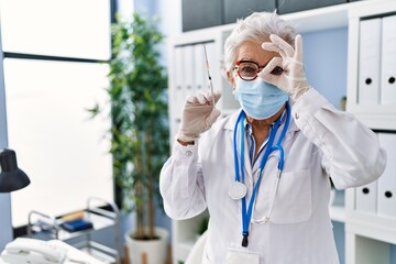 Senior woman with grey hair wearing doctor uniform and medical mask holding syringe smiling happy doing ok sign with hand on eye looking through fingers